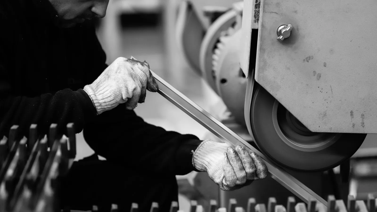 Mains gantées guidant une barre métallique contre une ponceuse à bande, en noir et blanc