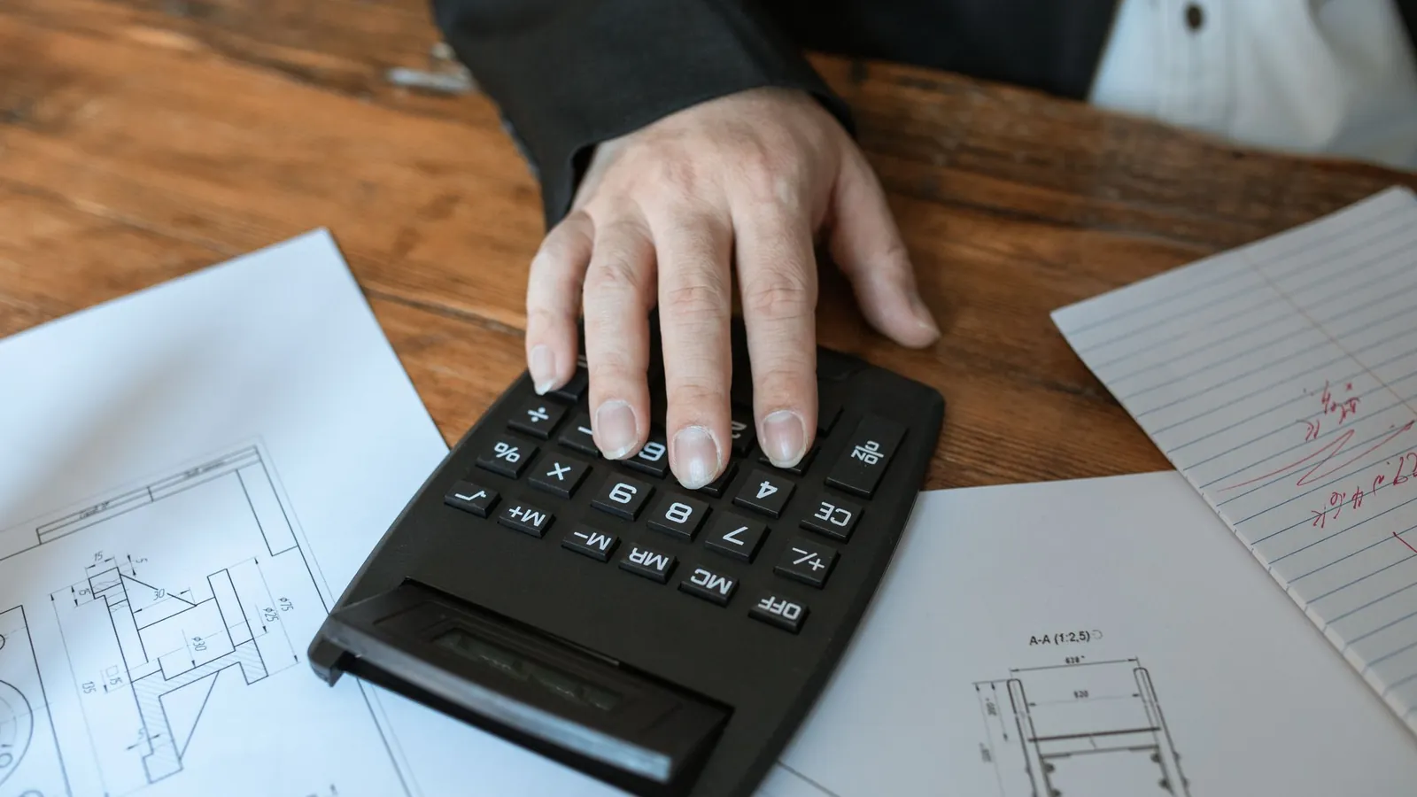 Main sur une calculatrice posée sur des plans techniques, pendant un chiffrage