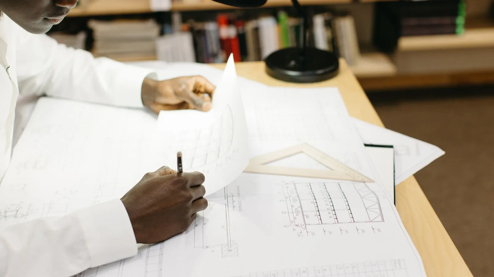 Mains annotant un plan technique au crayon sur un bureau, équerre posée à côté