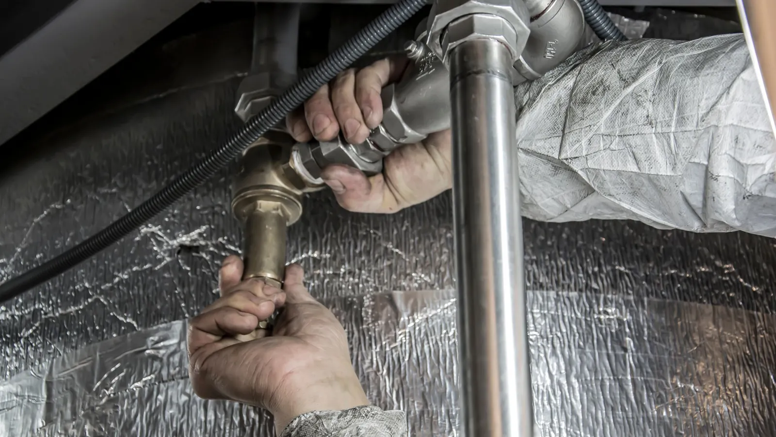 Mains serrant un raccord en laiton sur une tuyauterie en inox, sous une isolation