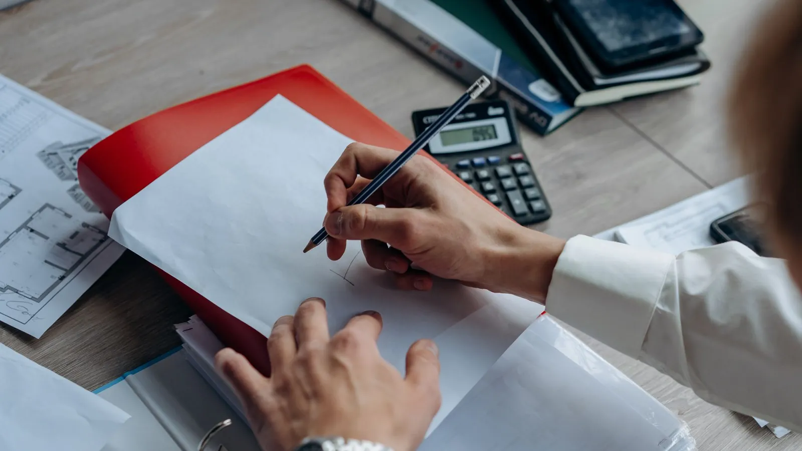 Mains rédigeant des notes sur une feuille, à côté d'une calculatrice, d'un classeur et de plans