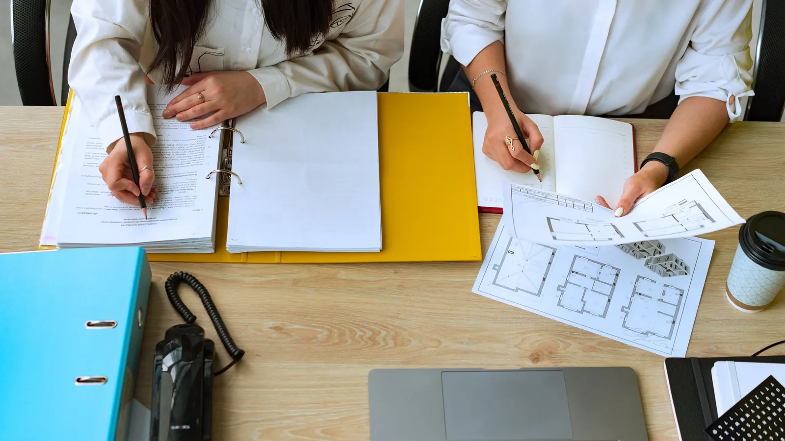 Deux personnes annotant des documents et des plans posés sur un bureau, entre classeurs et ordinateur