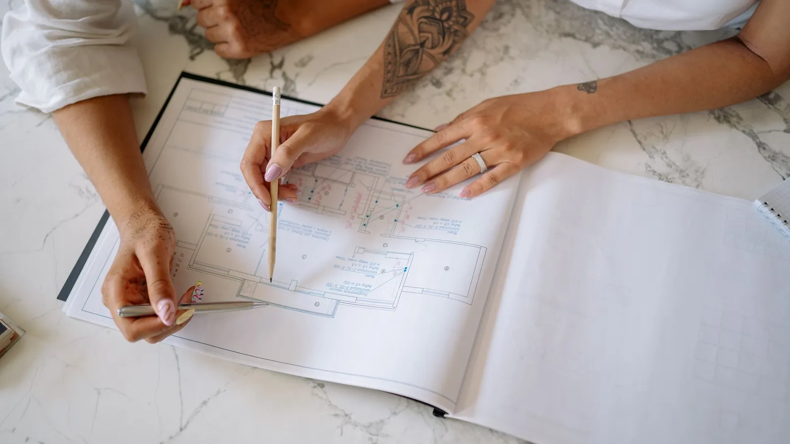Deux mains suivant au crayon les lignes d'un plan relié, posé sur une table en marbre