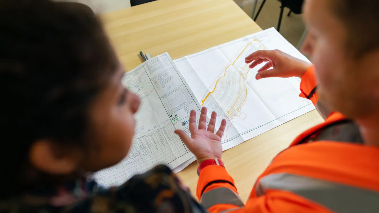 Deux interlocuteurs discutant autour d'un plan déplié sur une table, l'un en tenue de travail orange