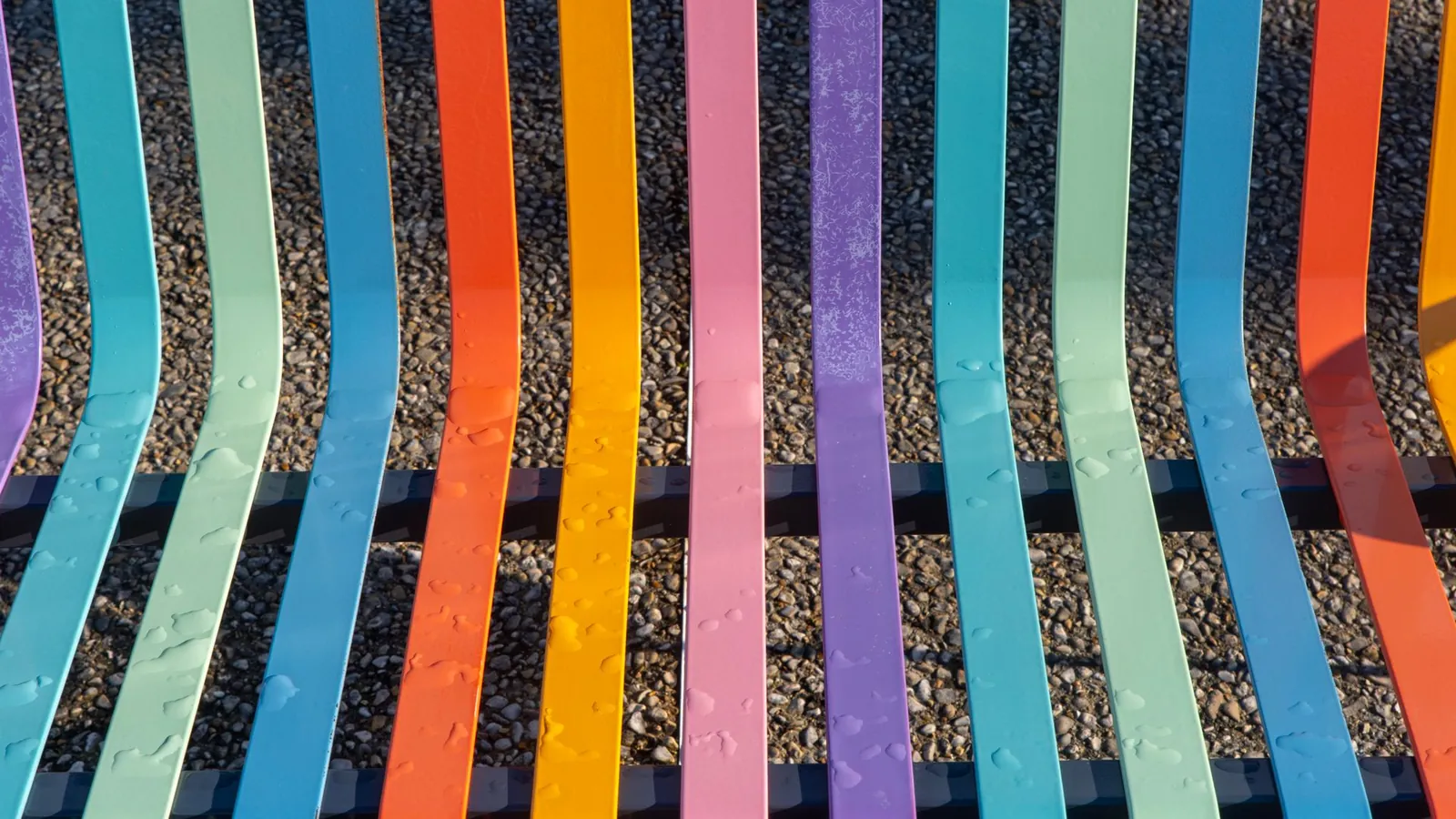 Banc public à lames métalliques laquées de couleurs vives, posé sur un sol de gravier