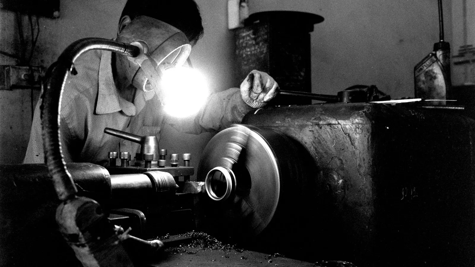 Tourneur protégé par des lunettes travaillant une pièce sur un tour, en noir et blanc