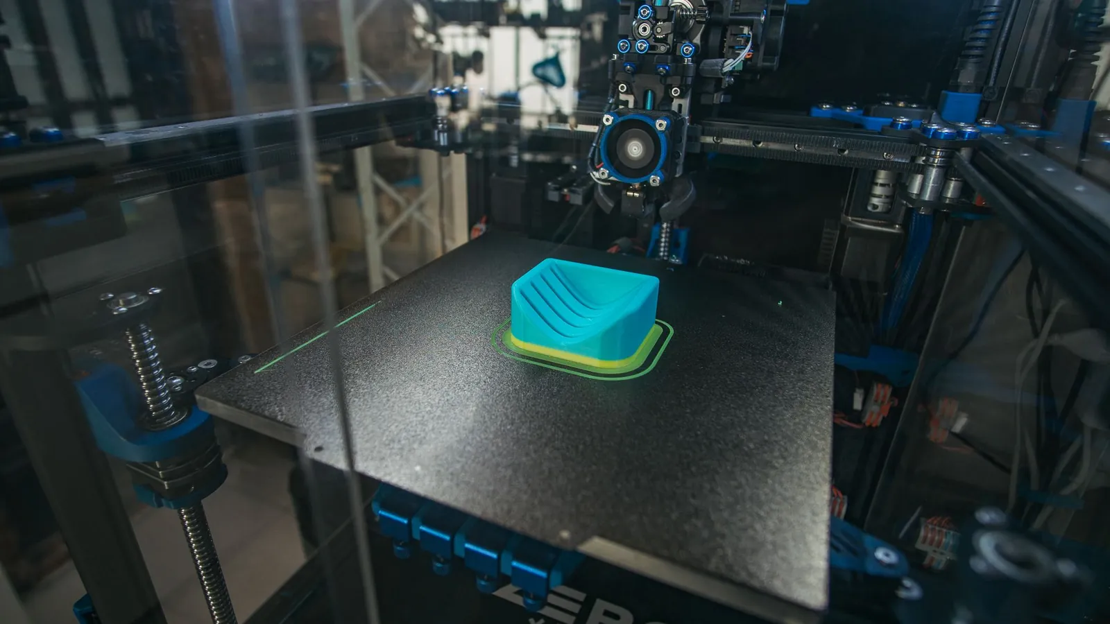 Imprimante 3D à dépôt de fil fondu en cours d'impression d'une pièce turquoise sur son plateau noir