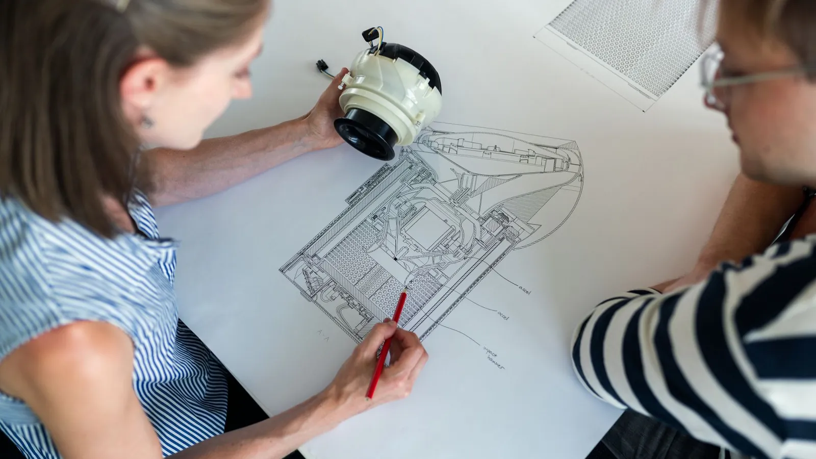 Annotation au crayon rouge d'un plan d'ensemble, le prototype de l'appareil tenu en main au-dessus du dessin