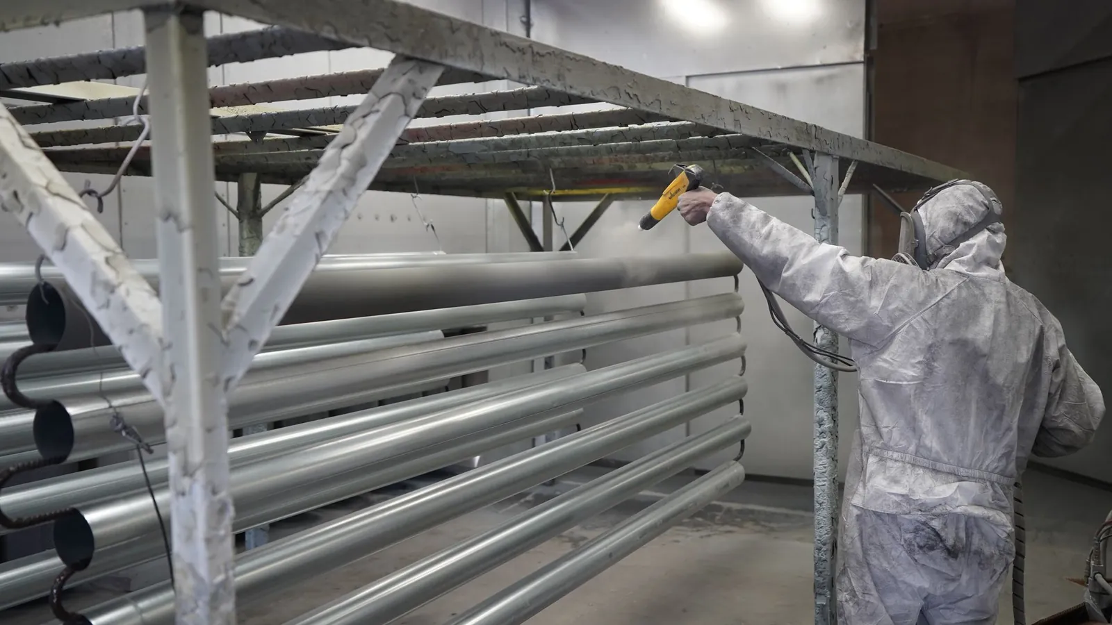 Opérateur en combinaison projetant de la peinture poudre sur des tubes métalliques suspendus dans une cabine