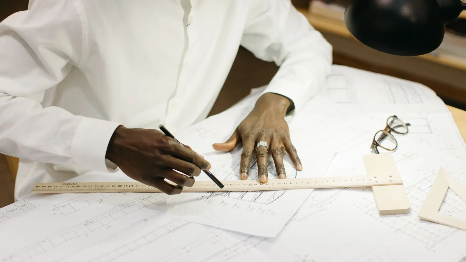 Mains traçant au crayon le long d'une règle posée sur des plans imprimés, lunettes et équerre sur la table
