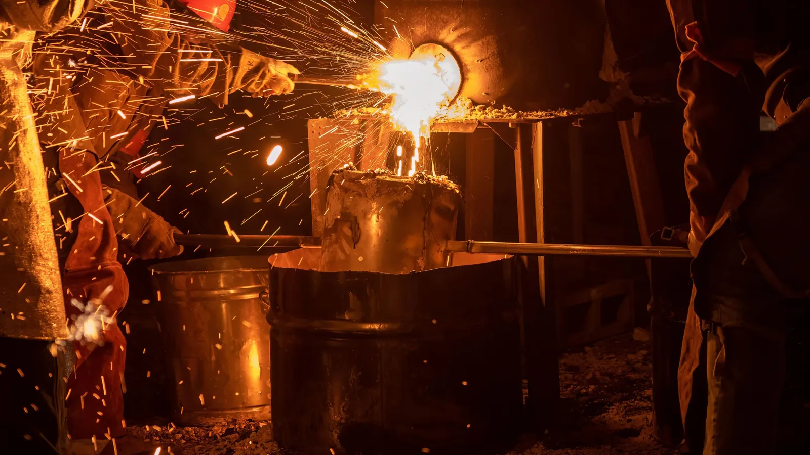 Métal en fusion s'écoulant du bec d'un four dans un creuset tenu par deux fondeurs, gerbes d'étincelles dans la nuit