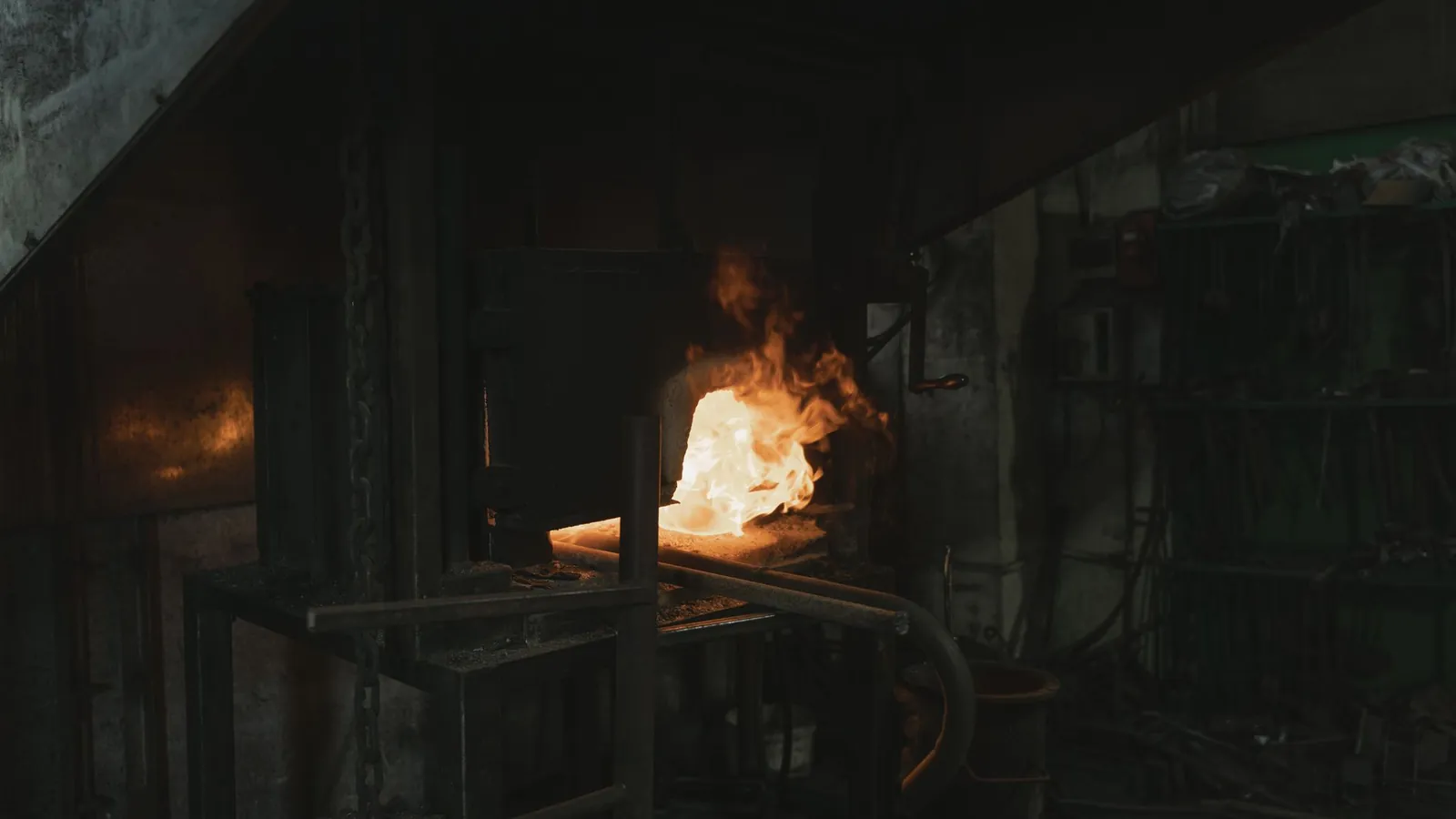 Four de forge allumé au fond d'un atelier sombre, flammes et lueur orangée à la bouche du four