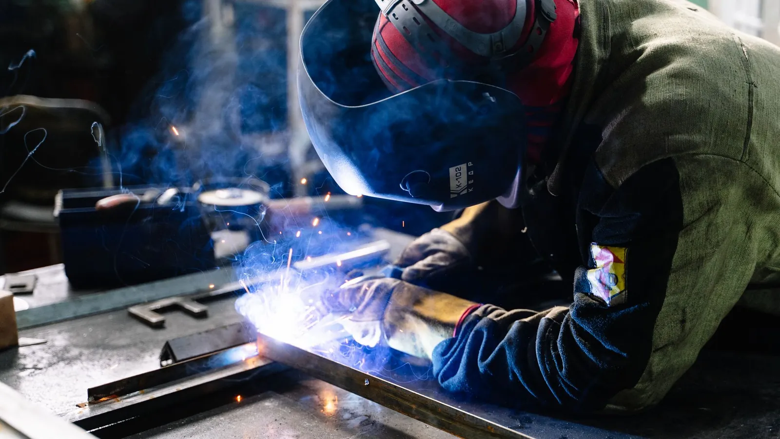 Soudeur casqué penché sur un cadre en profilés d'acier, arc bleu et fumée au point de soudure