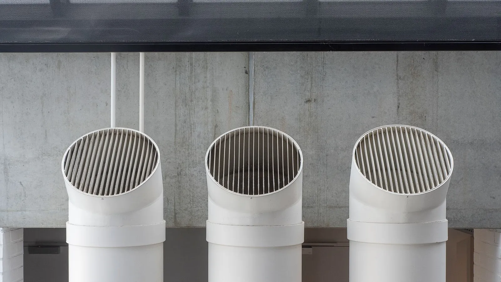 Trois tubes de ventilation en PVC blanc coudés, terminés par une grille, devant un mur en béton