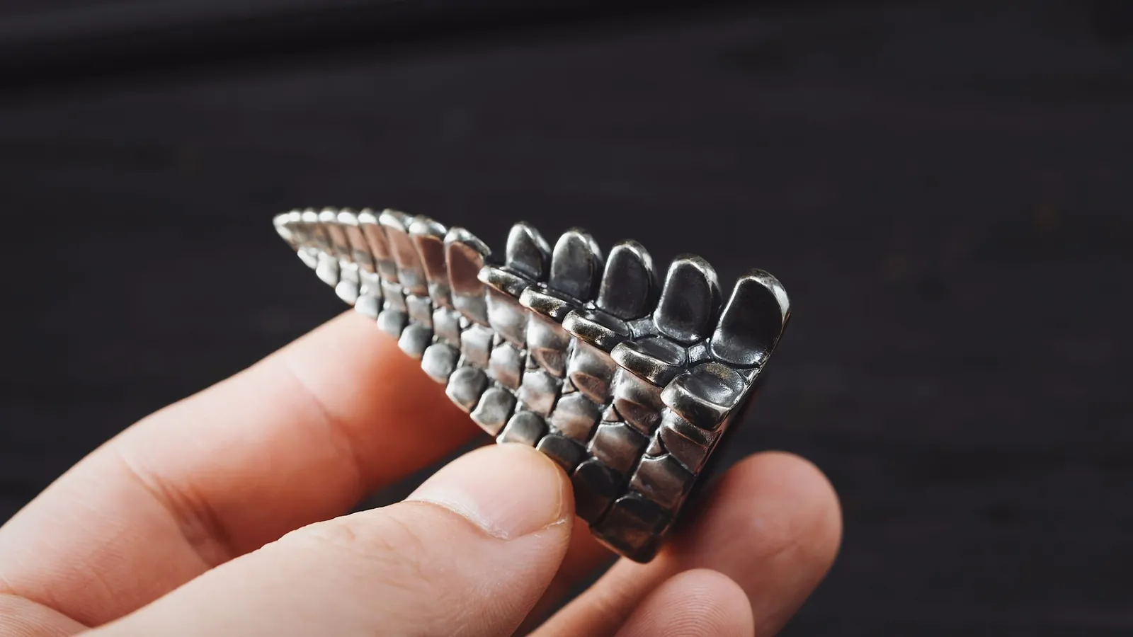 Petite pièce métallique coulée aux reliefs fins en écailles, tenue entre deux doigts sur fond sombre