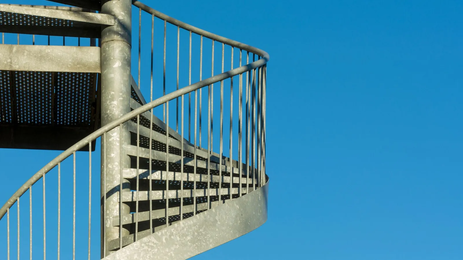 Escalier hélicoïdal extérieur en acier galvanisé, marches en caillebotis et garde-corps gris zinc sur ciel bleu