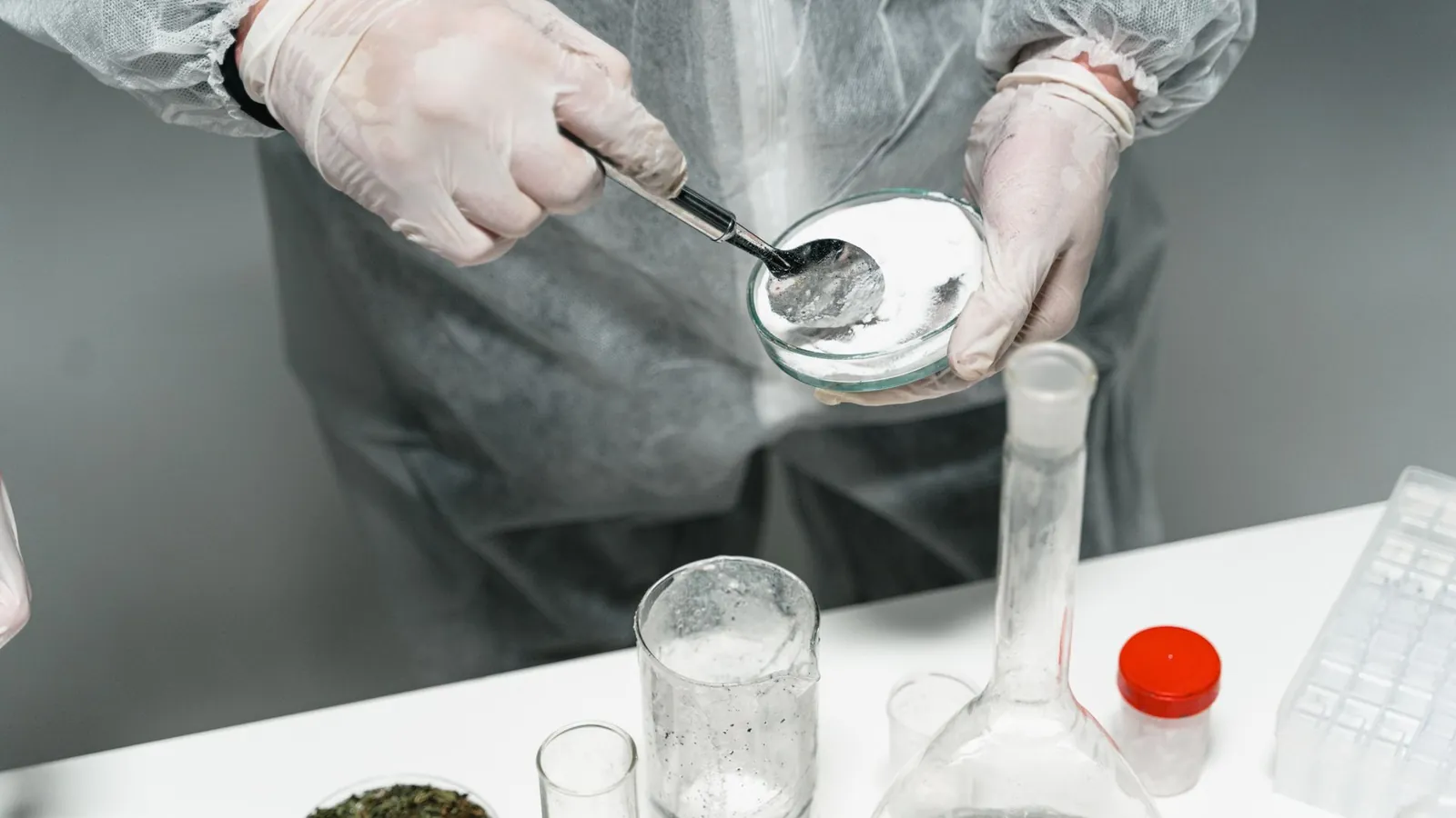 Opérateur ganté en blouse jetable prélevant une poudre fine à la spatule dans une coupelle de laboratoire