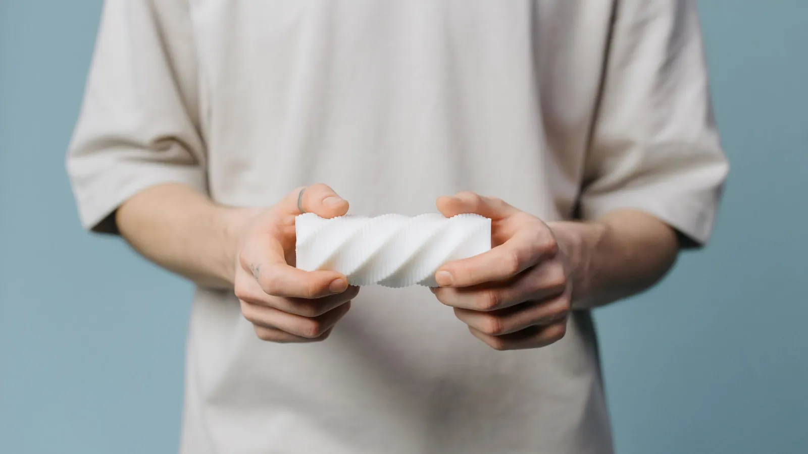 Mains tenant une pièce plastique blanche imprimée en 3D, de forme torsadée, sur fond bleu